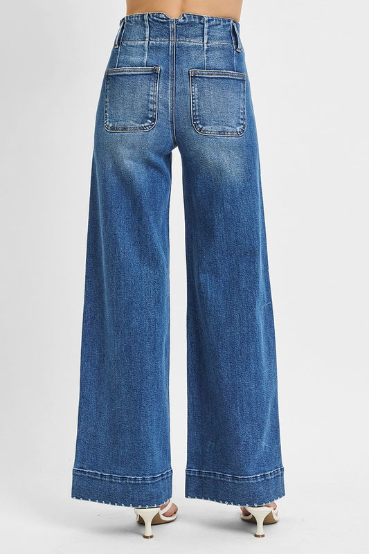 Model wearing high-rise wide-leg jeans in a medium blue wash featuring a double-button waist and raw hem cuffs, styled with white heeled sandals for a modern vintage look.