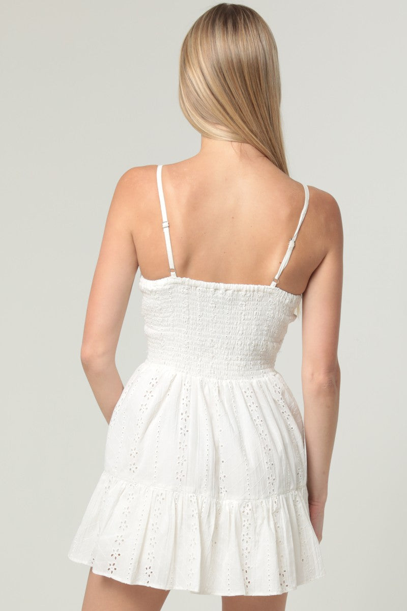 A romantic eyelet-embroidered romper featuring a twist-front bodice, cut-out waist, and flowing over-skirt.