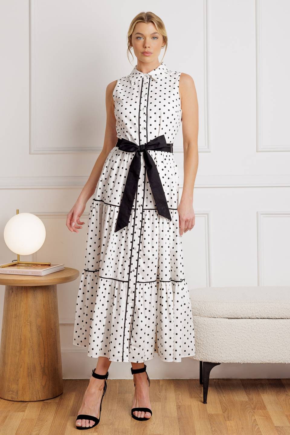 A white sleeveless maxi dress with black polka dots, a button-up bodice, tiered skirt, and a contrast black waist tie, styled with black strappy heels for a chic and timeless look.