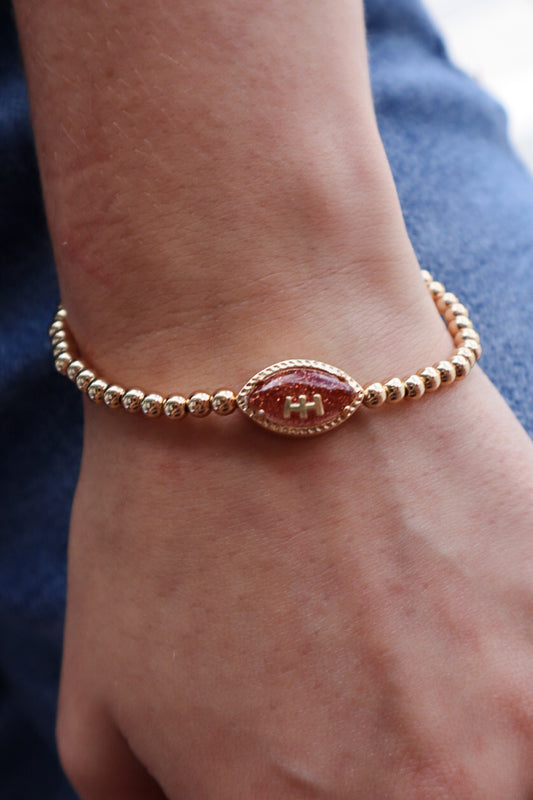 Gold beaded stretch bracelet with a sparkling football charm, displayed on a Pieces & Peaches Boutique card—perfect for football fans and game-day outfits.