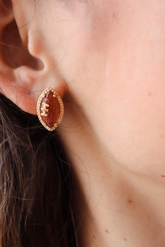 Football-shaped stud earrings with rhinestone accents on a gold-tone setting, displayed on a Pieces & Peaches Boutique card—perfect for game-day outfits and sports fans.