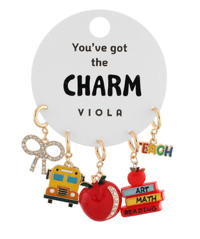 Set of five gold-toned clip-on charms on a white display card, featuring a glittery bow, a yellow school bus, a red apple with glitter, a stack of books labeled "READING," "MATH," and "ART" with a red apple and the word "TEACH" on top.