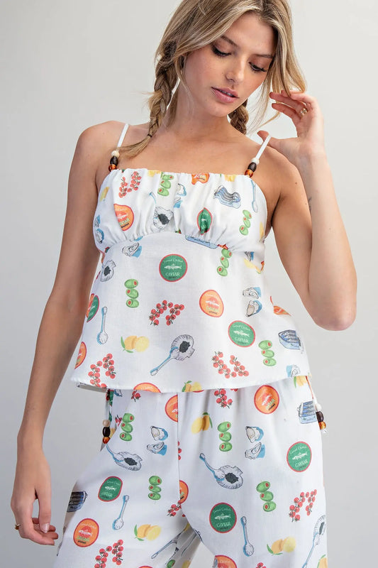 White Mediterranean-inspired food print babydoll top with adjustable beaded straps, gathered bust, and smocked back detail.