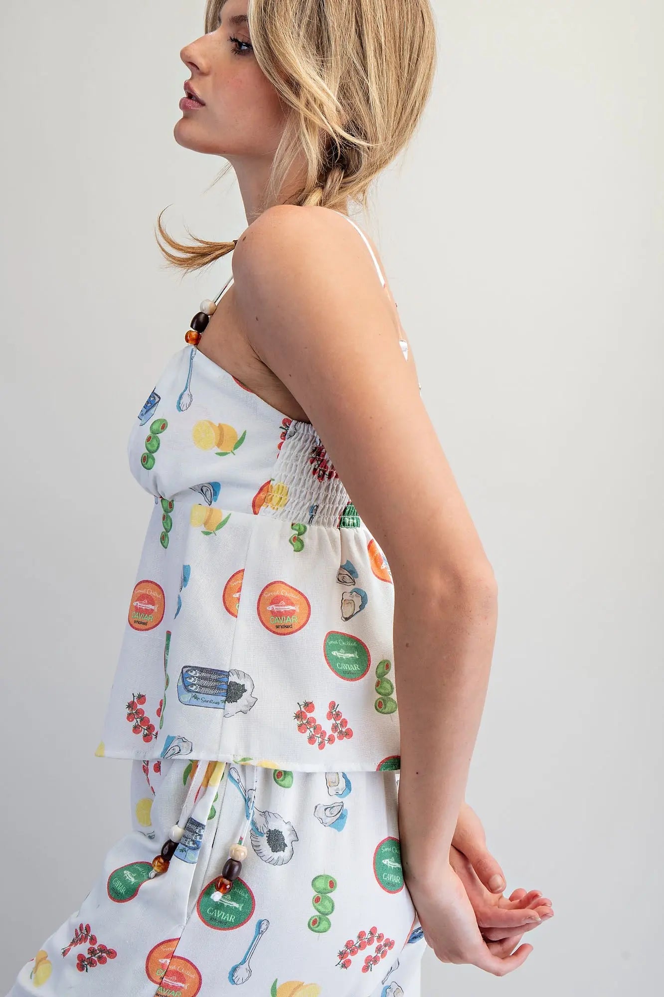 White Mediterranean-inspired food print babydoll top with adjustable beaded straps, gathered bust, and smocked back detail.