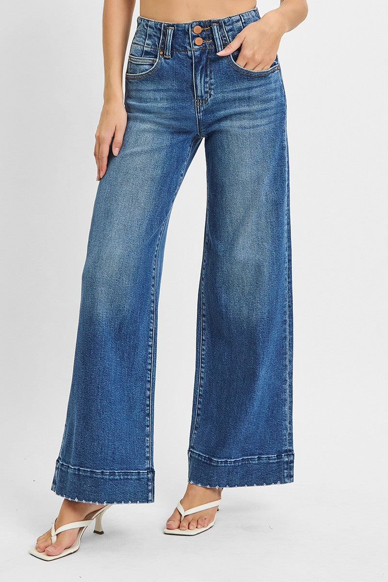 Model wearing high-rise wide-leg jeans in a medium blue wash featuring a double-button waist and raw hem cuffs, styled with white heeled sandals for a modern vintage look.