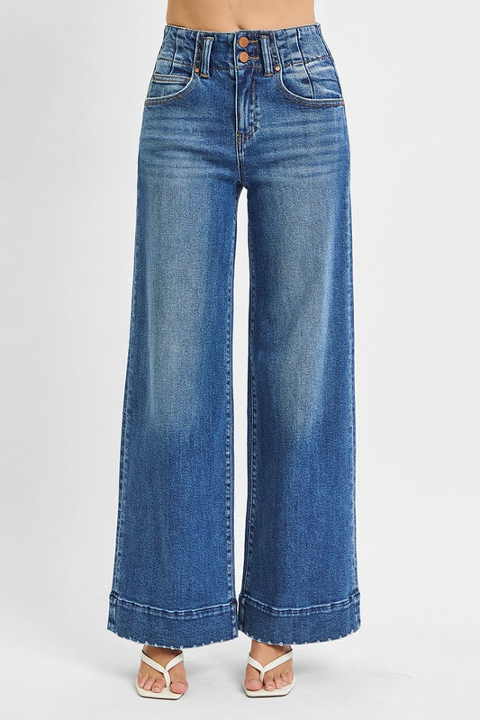 Model wearing high-rise wide-leg jeans in a medium blue wash featuring a double-button waist and raw hem cuffs, styled with white heeled sandals for a modern vintage look.