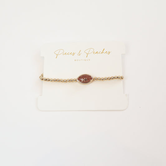 Gold beaded stretch bracelet with a sparkling football charm, displayed on a Pieces & Peaches Boutique card—perfect for football fans and game-day outfits.