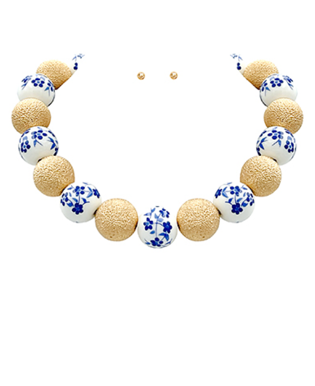 A vibrant statement necklace featuring a mix of large, spherical beads. Some beads are gold-toned with a textured, hammered finish, while others are white with intricate blue floral patterns reminiscent of porcelain. The necklace is designed to sit high on the neckline and has a simple gold-toned clasp.