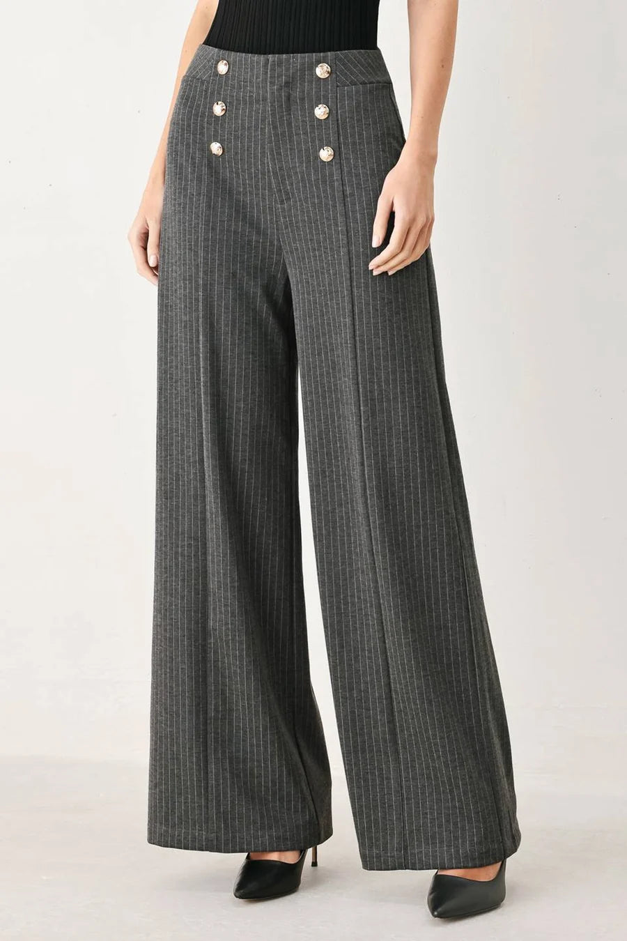 A model wearing high-waisted, wide-leg charcoal gray pinstriped trousers with decorative gold-tone buttons on the front waistband, paired with a black top and black heels.