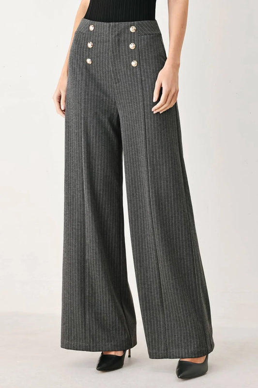 A model wearing high-waisted, wide-leg charcoal gray pinstriped trousers with decorative gold-tone buttons on the front waistband, paired with a black top and black heels.