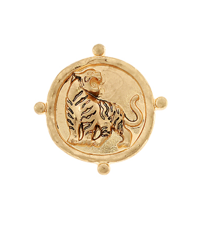 A round, gold-toned brooch featuring a raised, detailed depiction of a roaring or snarling tiger in the center. The tiger has distinct stripes and is depicted in a powerful, dynamic pose. The surrounding circular frame is also gold-toned and has small raised dots at the cardinal points, giving it a coin or medallion-like appearance.