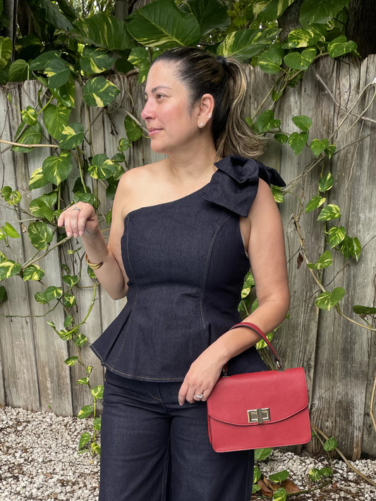 A woman wearing a dark navy one-shoulder peplum top and matching pants, holding a red handbag, standing in front of a wooden fence covered in green vines.