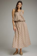 A woman modeling a sleeveless, midi-length maxi dress with a beige base and small dark floral print, set against a plain gray studio background.