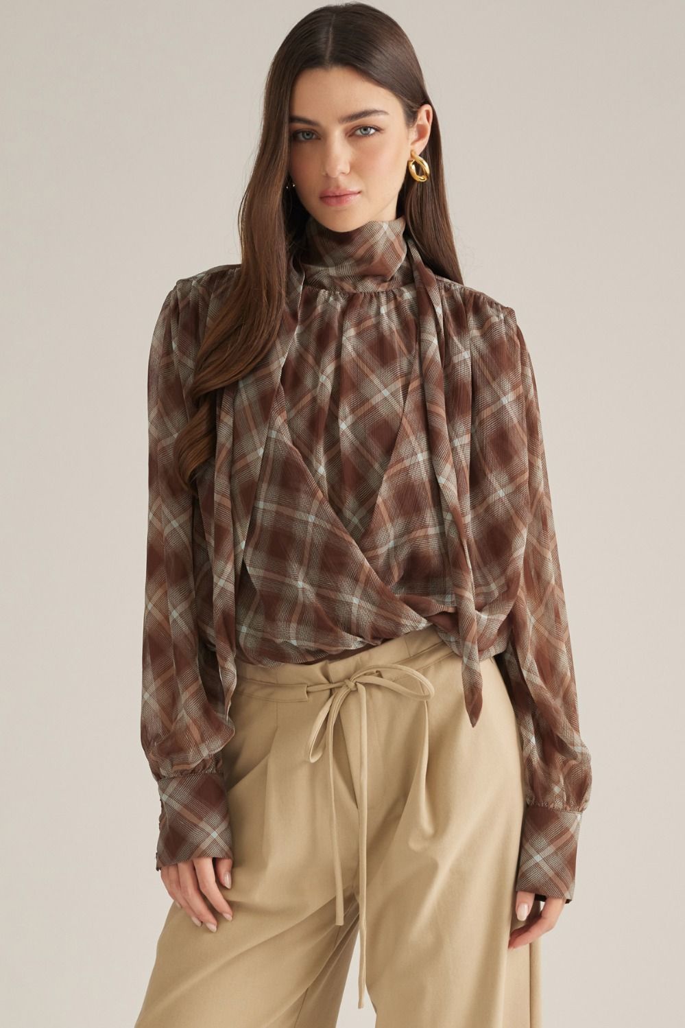 A sophisticated plaid blouse featuring a high neckline, elegant draped crossover front, self-tie back detail, and button cuffs.
