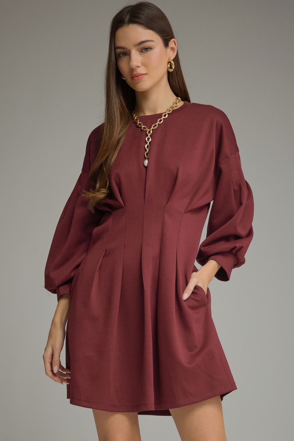 A timeless solid mini dress featuring a flattering pleated waist, classic round neckline, long sleeves, and convenient side pockets.

