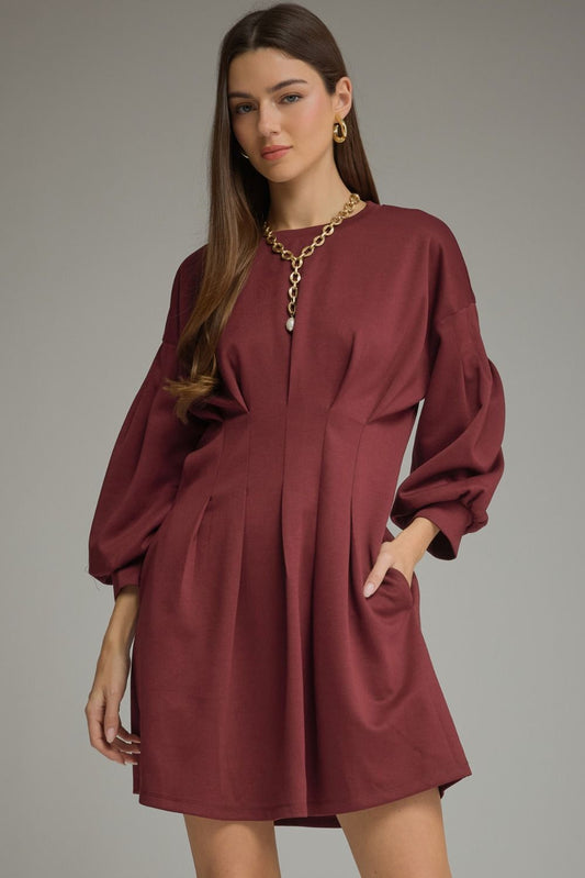 A timeless solid mini dress featuring a flattering pleated waist, classic round neckline, long sleeves, and convenient side pockets.

