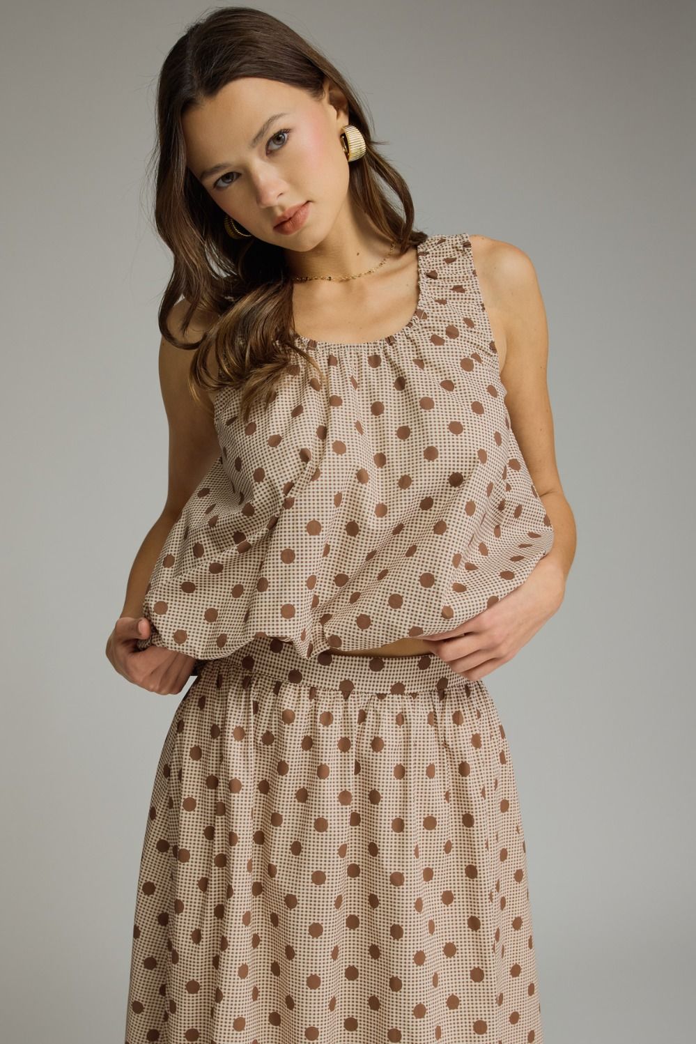 A woman modeling a sleeveless beige dress with a brown polka dot pattern against a solid gray studio background.